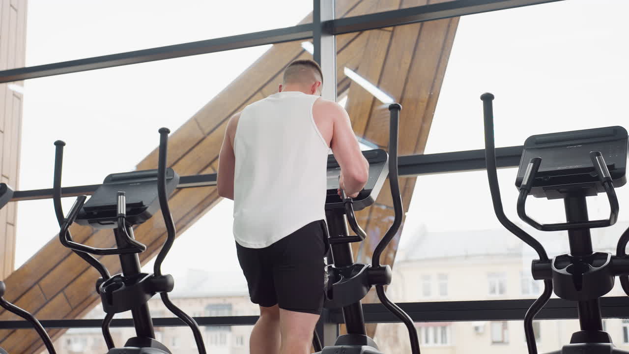Back view of trainer on elliptical by large glass window reflecting light, wearing white tank and black shorts, stepping on pedals and holding handles during cardio session in bright modern gym