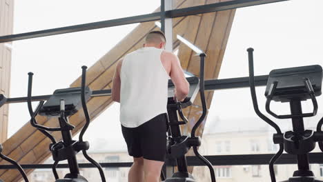 back view of trainer on elliptical by large glass window reflecting light, wearing white tank and black shorts, stepping on pedals and holding handles during cardio session in bright modern gym