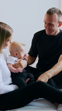 Beautiful Caucasian family sitting together on the big bed. Happy man plays with his baby and then kisses his wife. Vertical video