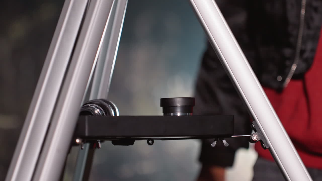 Geceleyin ellerin teleskop merceğini ve tripodunu ayarlaması, dikkatli odaklanma, metal tripod ayakları, küçük kontrol düğmeleri, görünen ceket kolu, yakındaki lens kapağı, loş soğuk ortam aydınlatması ve hazırlık yakın çekimi.