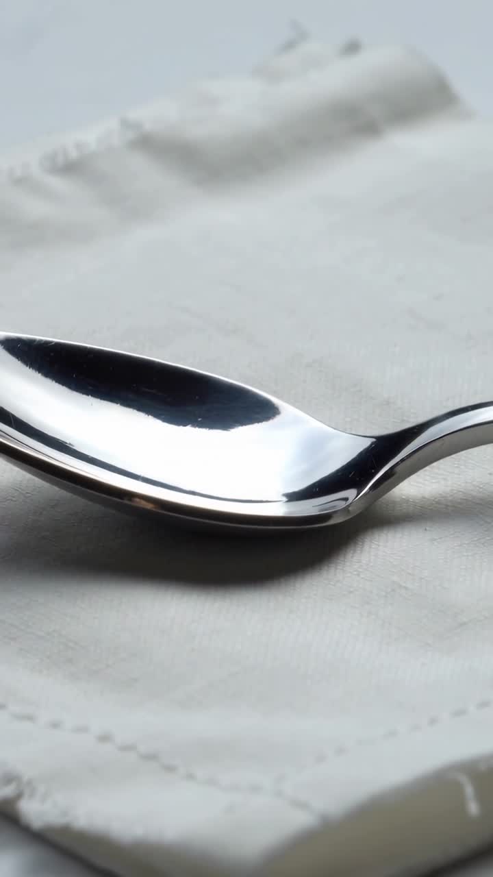 Vertical video: Pulling back camera revealing polished steel spoon on folded napkin, showing seam