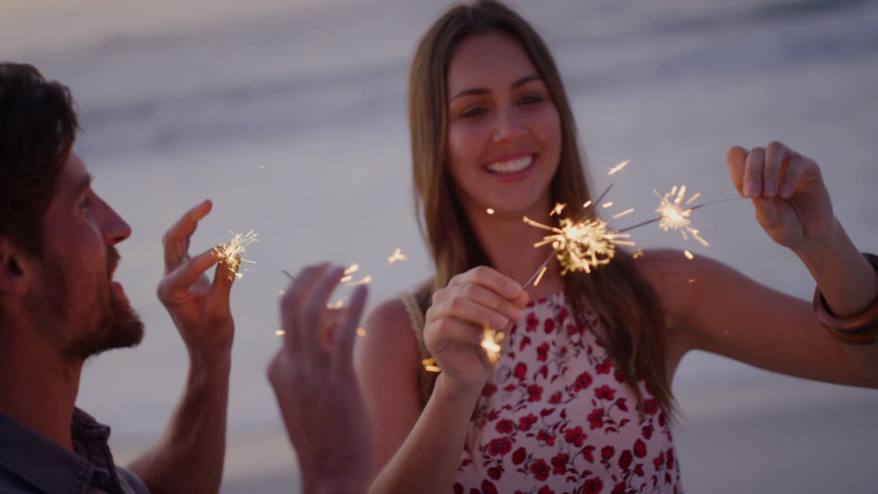 feliz pareja joven celebrando la víspera de año nuevo agitando bengalas en la playa al atardecer disfrutando juntos de una celebración romántica