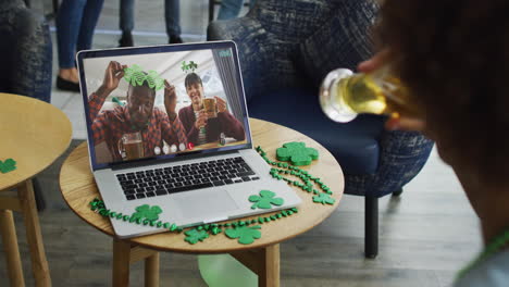 una pareja diversa sonriente con cerveza vistiendo artículos en forma de trébol en una videollamada en una computadora portátil