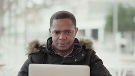 afro-american middle-aged man in black jacket with fur hood working on laptop outside