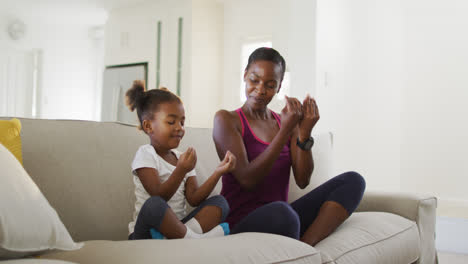 happy african american mother and daughter sitting on sofa doing yoga exercise at home