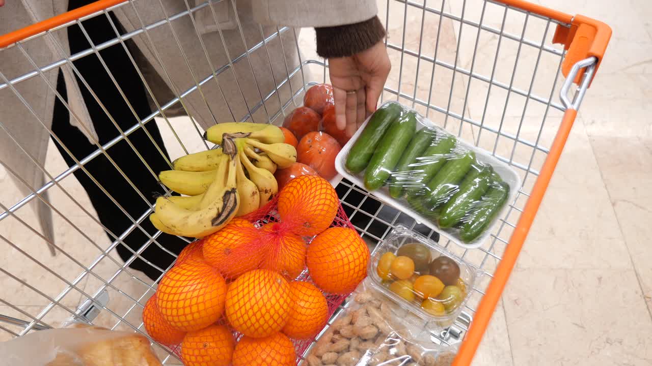 Person Placing Packaged Cucumbers into a Shopping Cart Full of Groceries