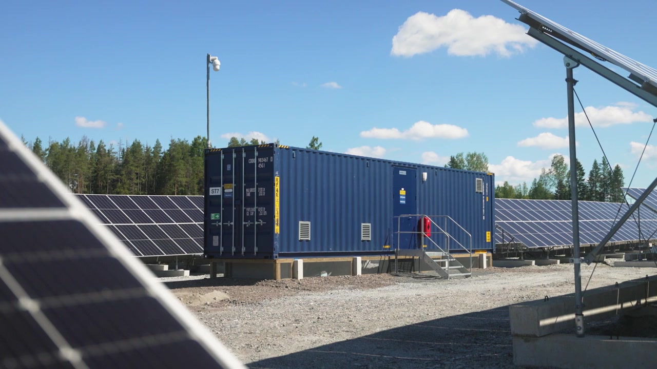 Kameraføring mod venstre afslører solpaneler i justerede rækker på en solcellepark med kontor i en shippingcontainer, fokus på ren vedvarende energi under solrig himmel.