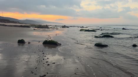 La-Escena-Cinematográfica-De-Caminar-Solo-Por-La-Playa-Cerca-De-Las-Olas-Del-Mar-Que-Salpican-La-Orilla-Al-Atardecer,-Revelando-La-Superficie-Del-Océano