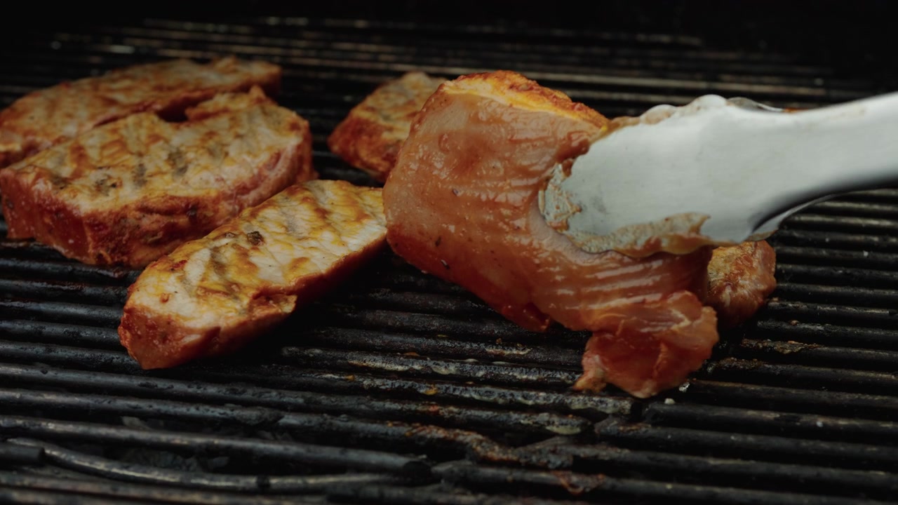 Des pinces argentées retournent d'épais morceaux de porc assaisonnés qui cuisent sur des barres de gril chaudes, avec un brunissement visible et de la fumée.