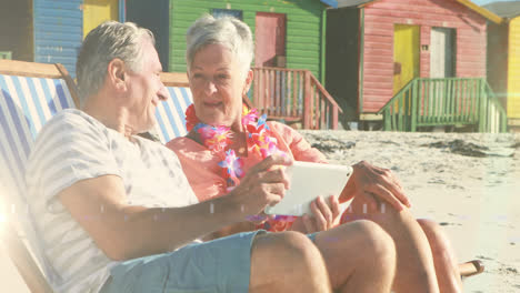 Animación-De-Luz-Brillante-Sobre-El-Retrato-De-Una-Feliz-Pareja-De-Ancianos-Usando-Una-Tableta-En-Tumbonas-En-La-Playa.