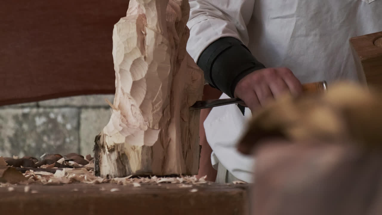 Static shot with a blurred object in the foreground as an artisan carves a log with chisel and mallet in the background; tools and wood shavings scattered across the table