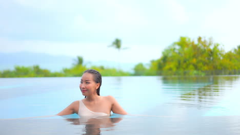 asian woman standing half submerged in the swimming pool water, looking around , with tropical out of focus background behind her