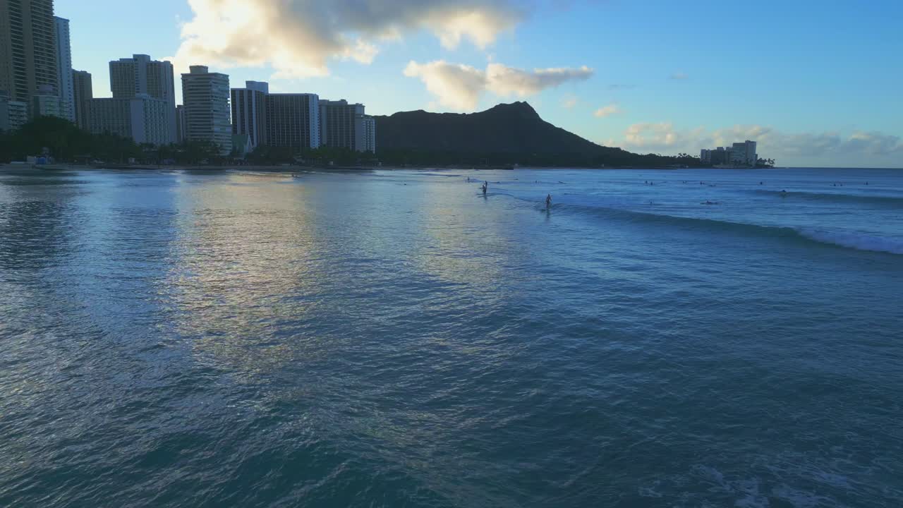 Dolly in Aerial drone shot in slow motion apporaching surfers riding waves in Waikiki Beach, Hawaii, at sunset