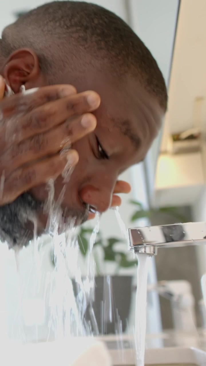 Vertical video: Chrome faucet running as African American man lathering and rinsing beard at sink