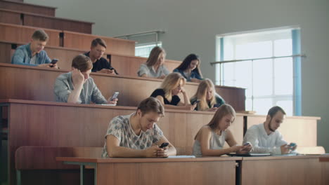 Eine-Gruppe-Von-Männern-Und-Frauen-In-Einem-Großen-Universitätspublikum-Schaut-Schweigend-Auf-Die-Bildschirme-Ihrer-Smartphones-Und-Schreibt-Chatnachrichten.-Jeder-Einzelne-Schaut-Auf-Den-Bildschirm-Seines-Geräts,-Ohne-Auf-Das-Geschehen-In-Der-Realen-Welt-Zu-Achten.