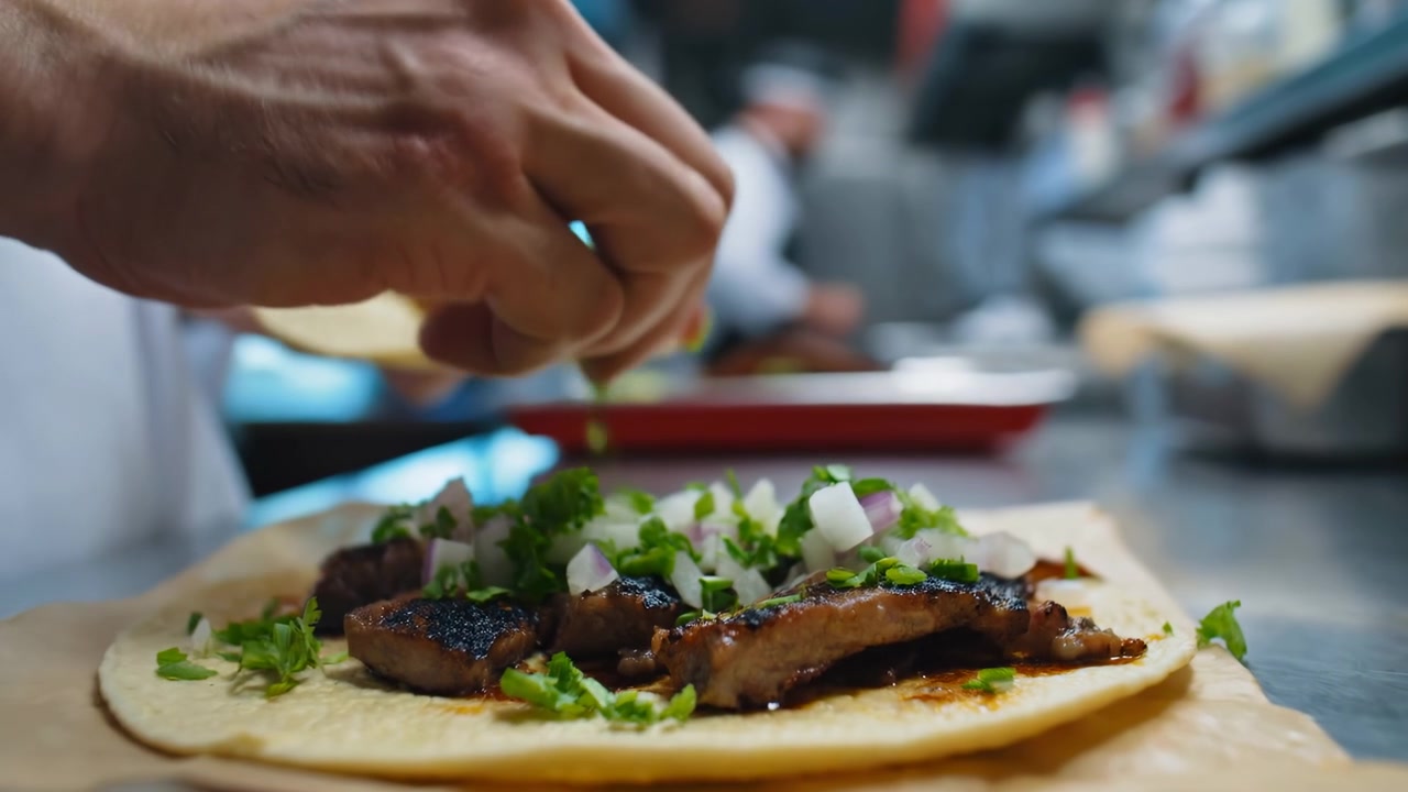 Nærbillede af kokkehænder, der samler en grillet taco på en varm tortilla, topper med hakket løg og koriander, travl atmosfære.