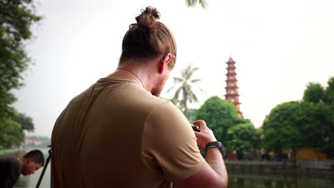 travel photographer setting up camera on tripod with pagoda of tran quoc temple in blurry distance. hanoi, vietnam. medium shot