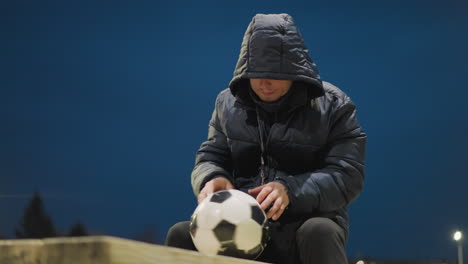 un hombre sentado en un banco con una pelota de fútbol entre las piernas, devuelve su teléfono a su bolsillo, y comienza a girar la pelota de una manera lúdica