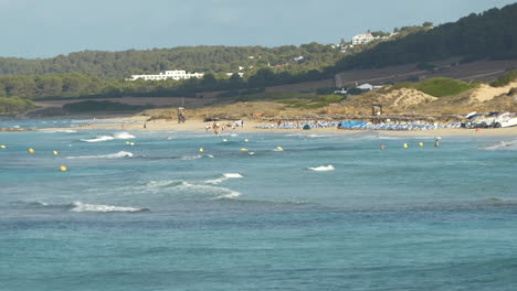 una vista lejana de una playa de menorca con olas pequeñas y turistas nadando irreconocibles