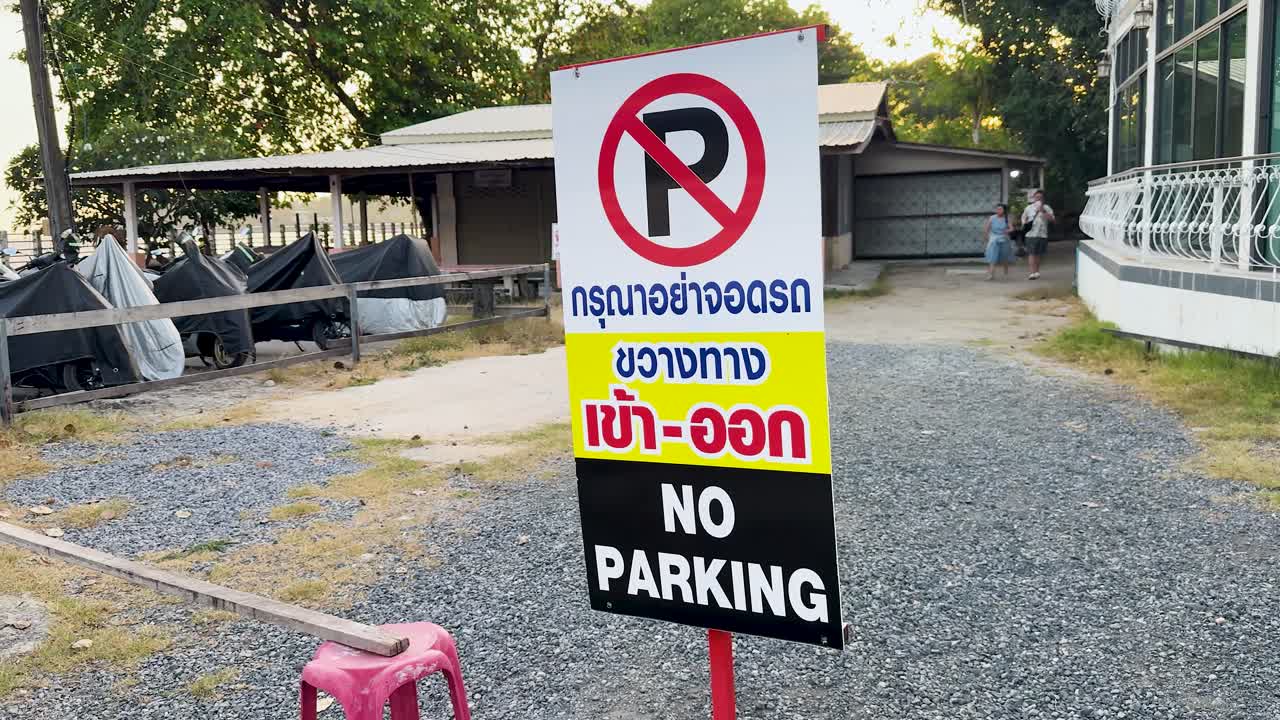 Camera moves past bilingual no parking sign in outdoor gravel lot, daylight, urban Thailand setting