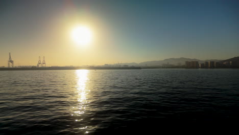 a serene sunset over the water at malaga port, with the sun casting a golden reflection on the sea and cranes visible in the distance