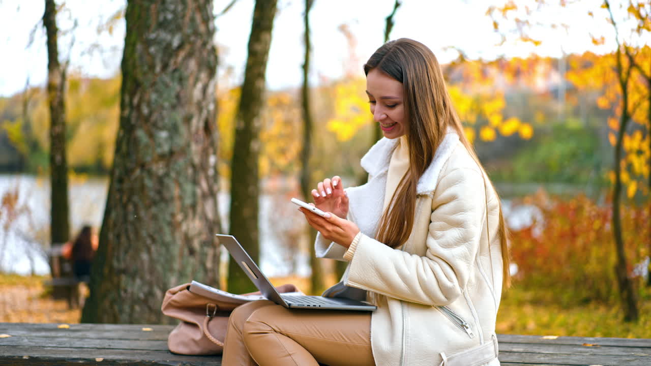 Happy smiling young woman using gadgets resting in the nature. Lady looks at her phone smiling and having laptop on her laps. Blurred nature autumn backdrop.