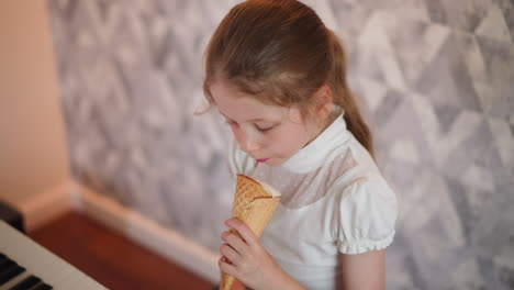 cute child in white top enjoys ice cream cone while casually practicing piano indoors, focused and happy, sitting beside modern keyboard with soft patterned gray wall in background