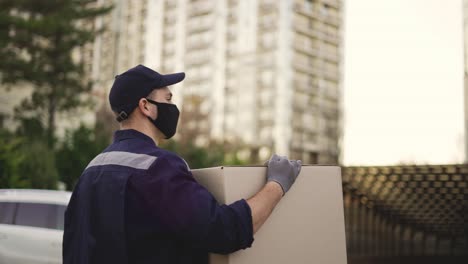 Zusteller-In-Uniform,-Maske-Und-Handschuhen,-Der-Ein-Großes-Kartonpaket-Im-Freien-Trägt