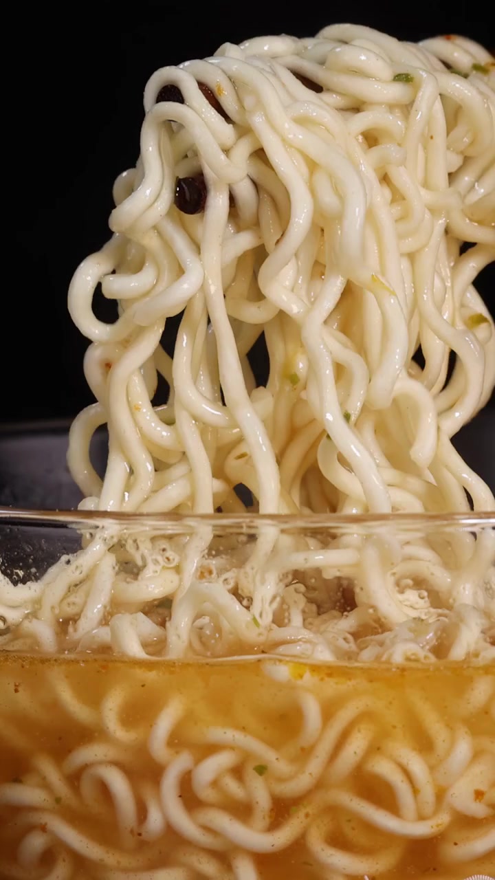 黒い背景とドラマチックなスタジオ照明の下、箸が熱いスープで満たされた透明なガラスの丼から調理されたインスタント麺をつかんで持ち上げる。