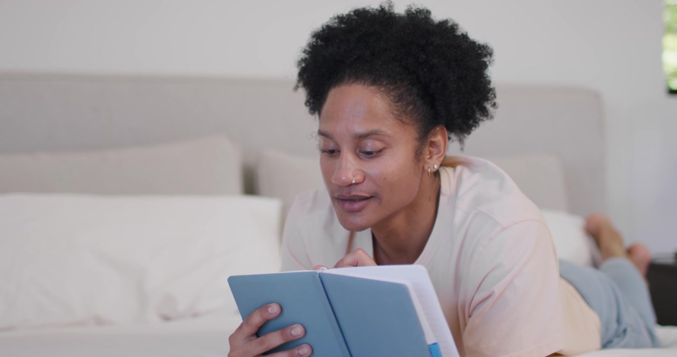 African American man lying on bed holding blue notebook, tapping pen to lips, reading and studying