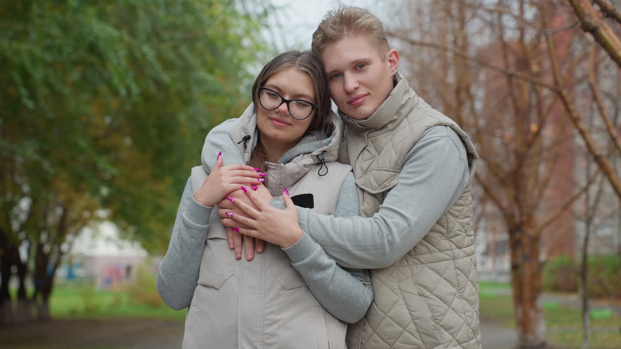Beautiful moment of lovely lovers warmly holding each other outdoors as man gently rests head on her head, both dressed in light jackets with soft expressions, surrounded by trees and blur background