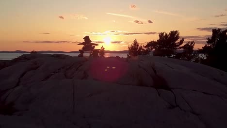 reveal of inukshuk on rocky pine tree island at sunset, drone aerial wide dolly out