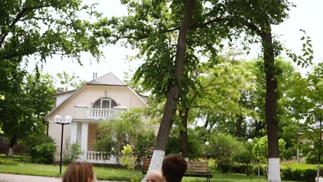 Schönes-Ferienhaus,-Greet-Street.-Junge-Dreiköpfige-Familie-Ruht-Auf-Einem-Plaid-Im-Park.-Bäume-Herum.-Kaukasischer-Mann-Wirft-Seinen-Kleinen-Sohn-In-Die-Luft.-Glückliche-Familie.-Zeitlupe