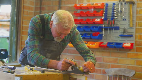 senior male carpenter wearing overalls in garage workshop planing piece of wood