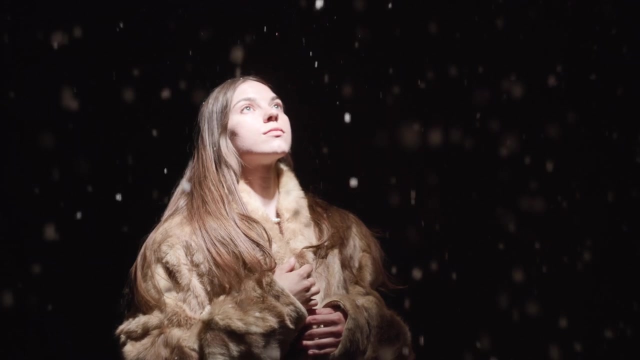 Una giovane donna con un cappotto di pelliccia testurizzato sta sotto un caldo riflettore su un palco scuro, creando un ritratto di moda drammatico con uno stile elegante