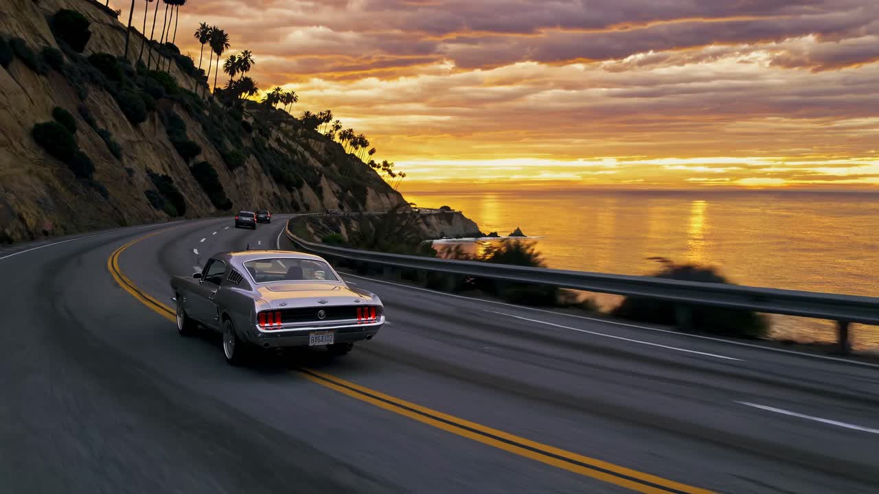 Un'auto d'epoca percorre un'autostrada costiera al tramonto, ripresa da un angolo posteriore basso.