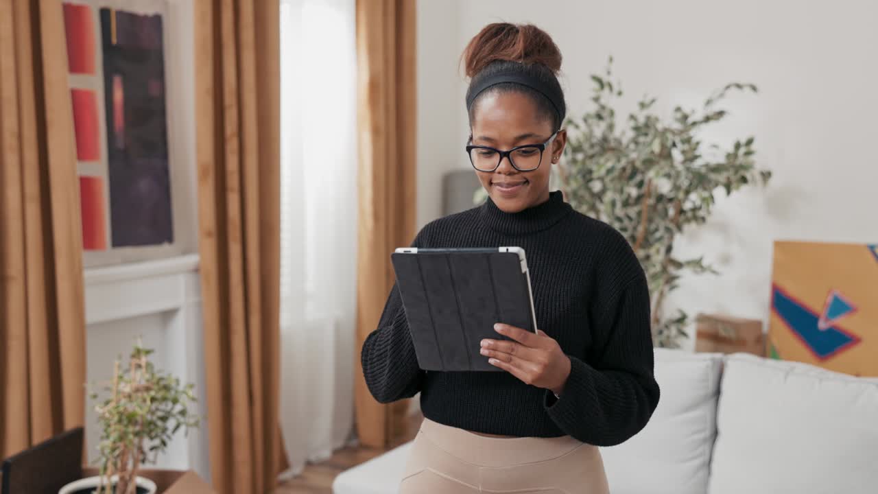 Smiling girl stands in the middle of the apartment about renovation, new house, moving boxes stand in the living room, the woman holds a tablet in hands, buys online furniture for flat