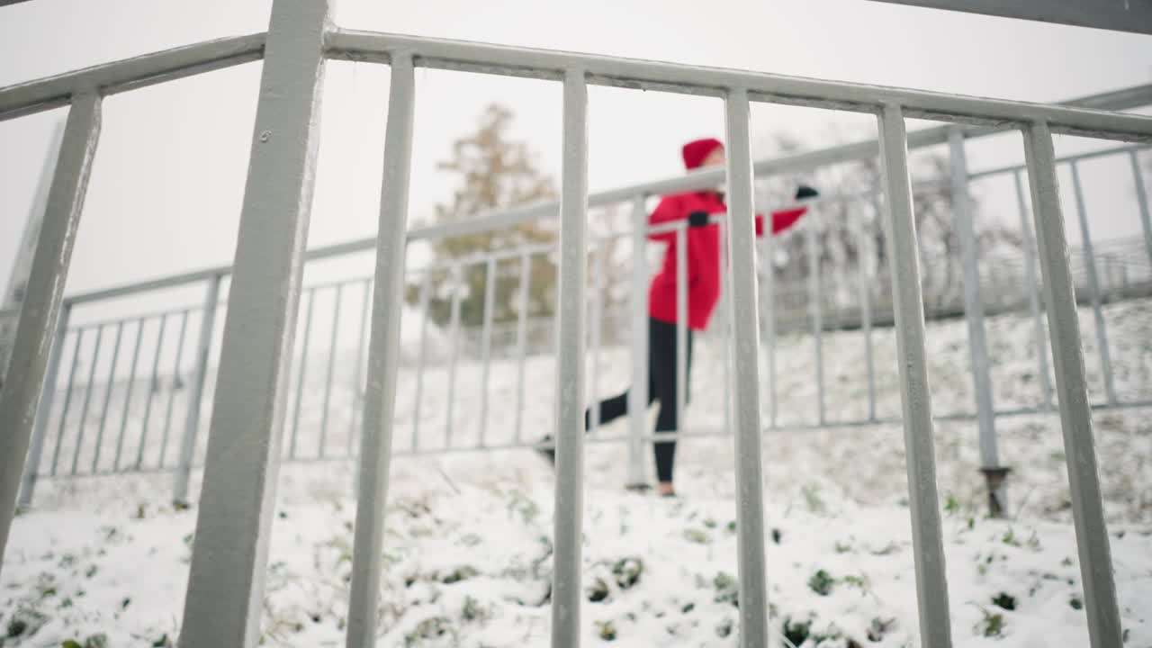 Close up of iron rail covered with snow frost, snowy ground, and blurry background of woman working out by stretching her leg back and forth, surrounded by trees in a foggy winter atmosphere