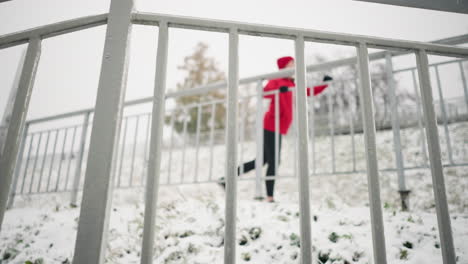 close up of iron rail covered with snow frost, snowy ground, and blurry background of woman working out by stretching her leg back and forth, surrounded by trees in a foggy winter atmosphere