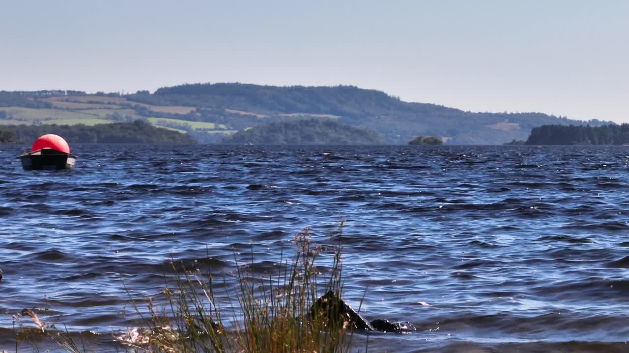 Niedrige Perspektive am Ufer des Loch Lomond an einem ruhigen, sonnigen Tag mit anrollenden Wellen und einem kleinen Boot.