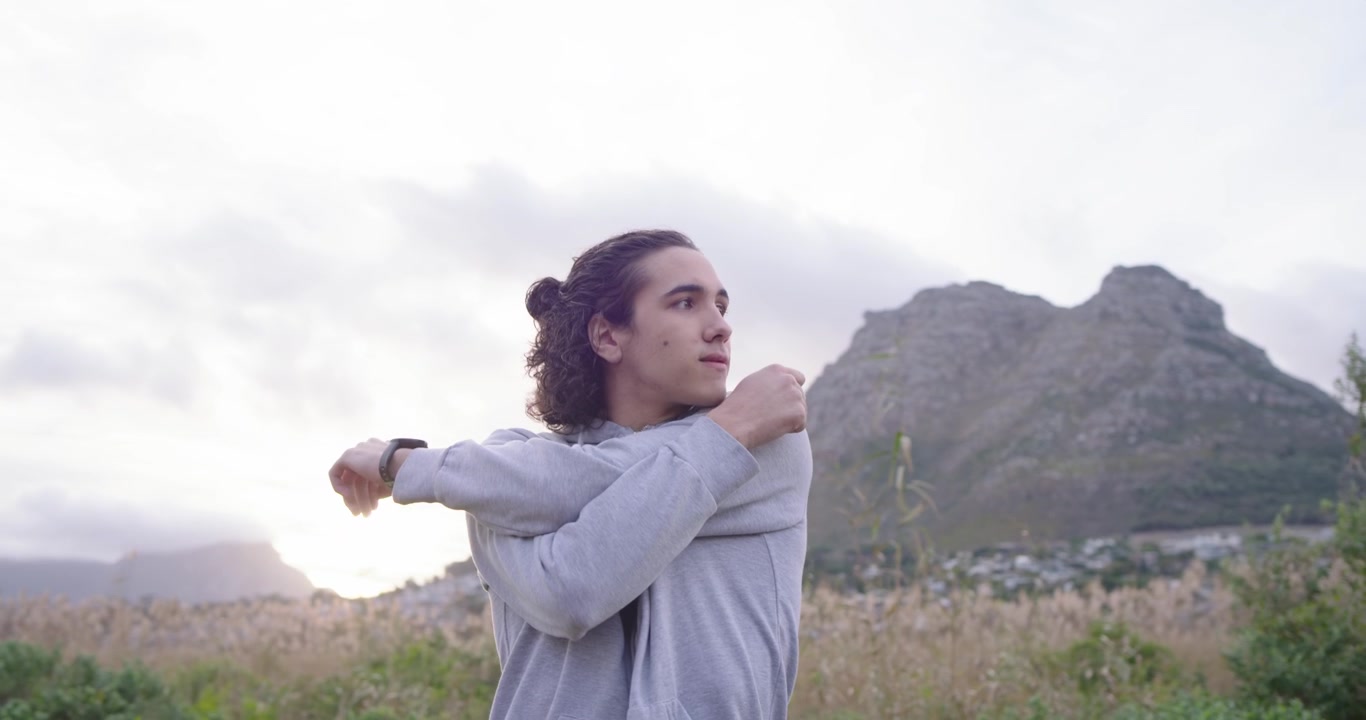 African man doing shoulder stretches before run in meadow, wearing gray hoodie with tracker