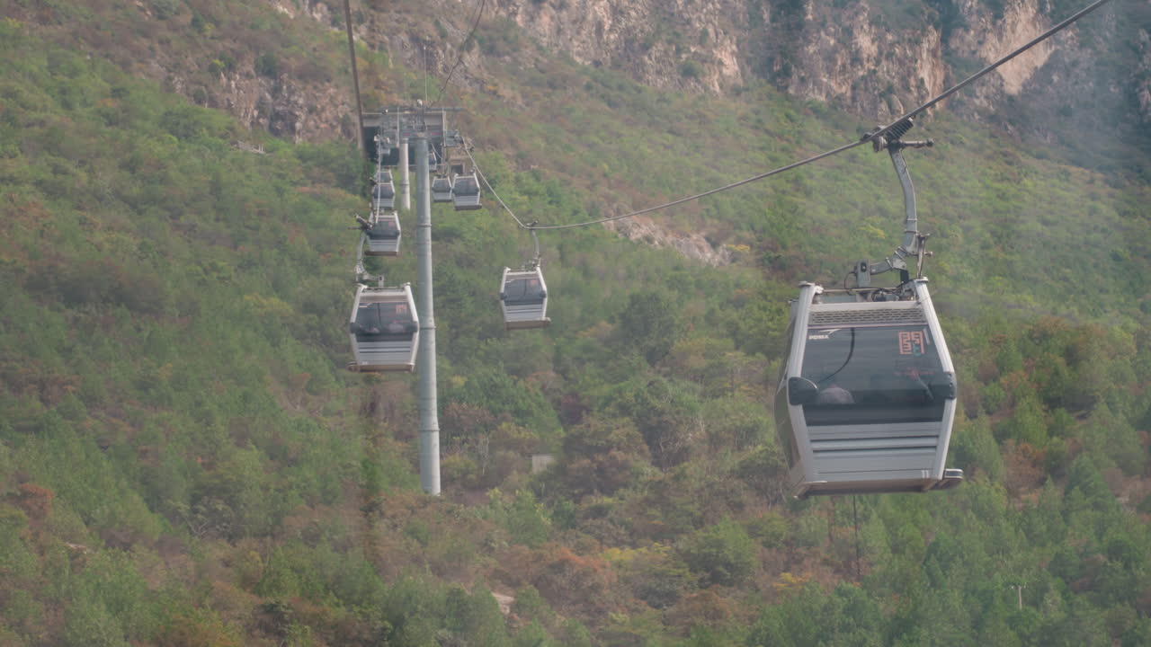Una telecabina attraversa una valle verde e aspra, una navetta per pendolari ronza tra le torri, un tecnico monitora la tensione dei cavi, una guida indica formazioni rocciose sulle pareti del dirupo, movimento costante con precisione industriale.