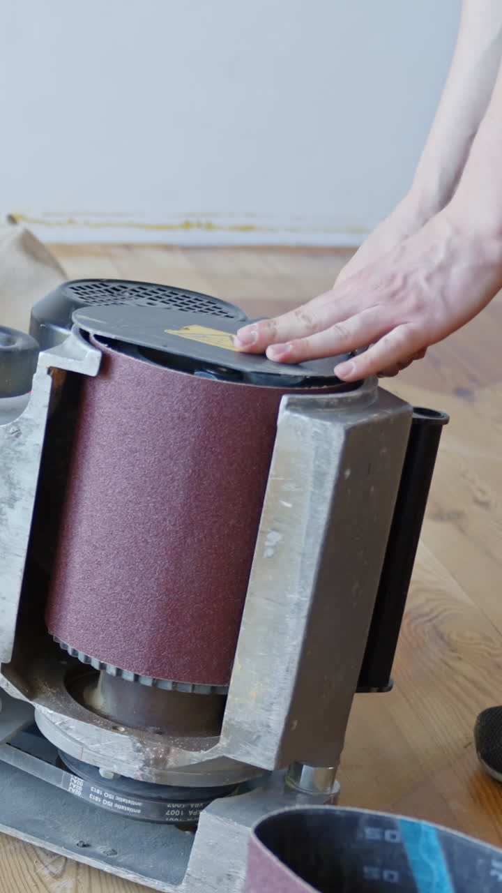 Vertical close-up of person hands finishing the process of installing a fresh sandpaper roll onto professional floor sanding machine, focusing on precise alignment and secure fit for wood renovation.