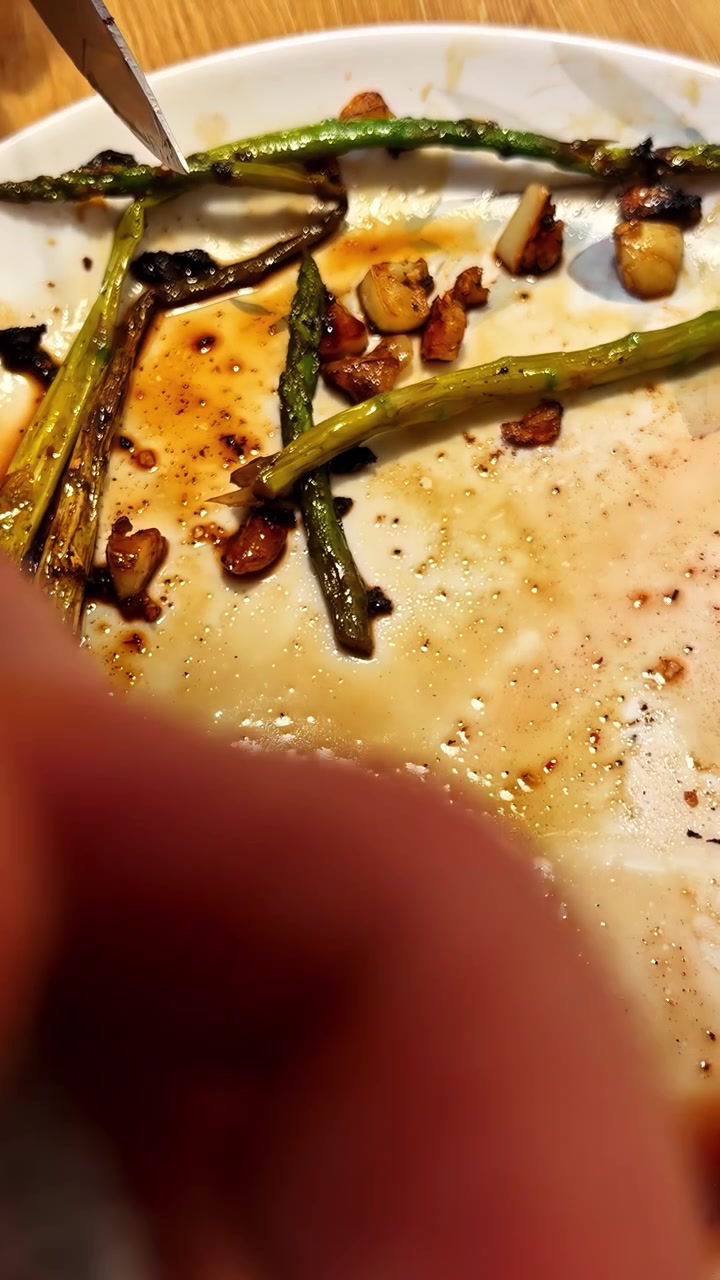 A hand uses a fork and knife to move roasted asparagus and garlic pieces on a white plate with sauce remnants on a wooden table. Vertical video, ideal for social media.