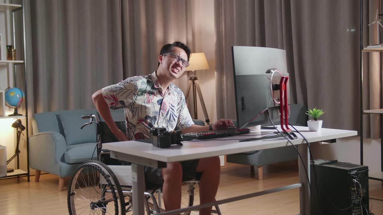 체어 에 앉아 있는 아시아인 남자 가 집 에 있는 카메라  에 있는 데스크 으로 비디오 를 편집 하는 동안 허리 통증 을 겪고 있다
