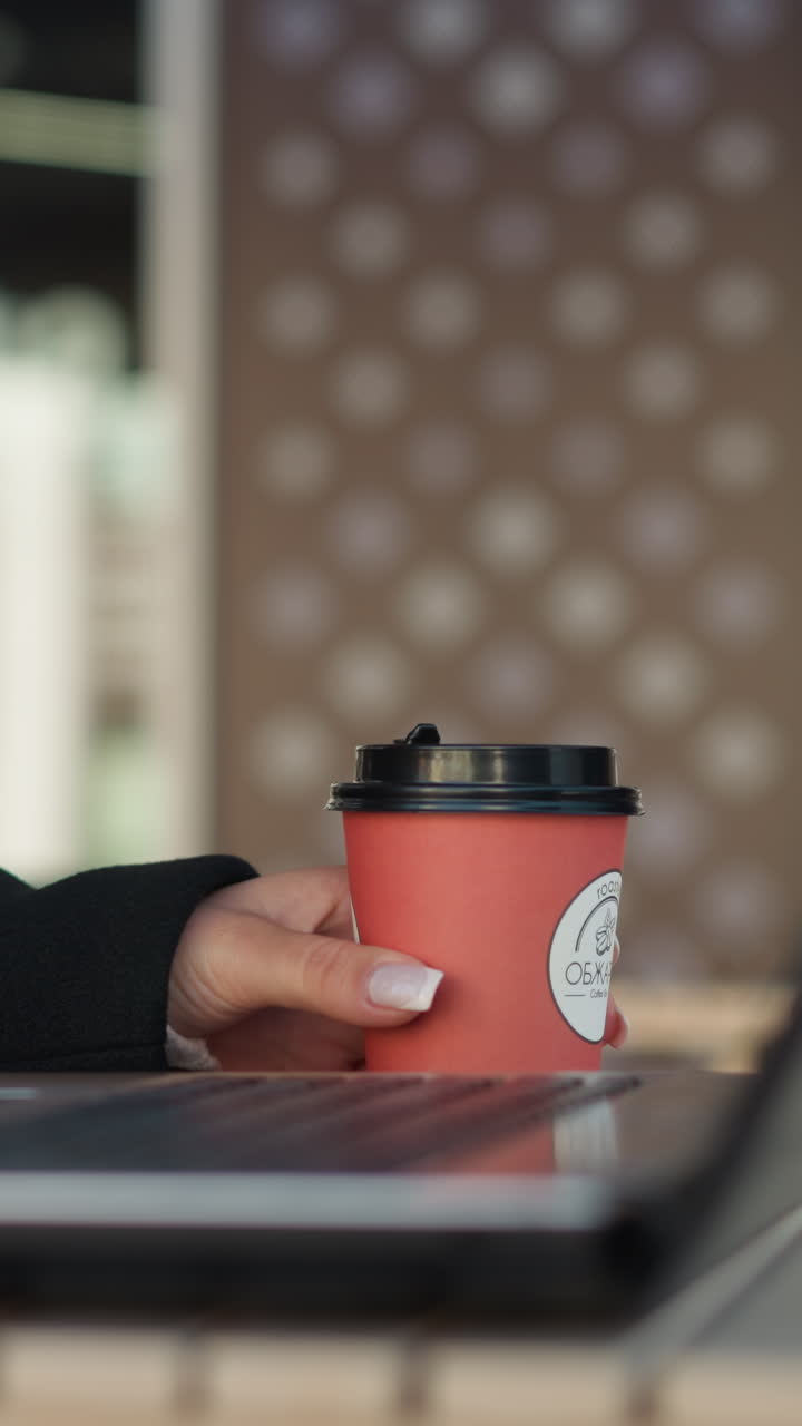vista lateral de la computadora portátil en la mesa con un teclado visible, persona que sostiene una taza de café cerca de la laptop, la deja caer en la mesa y comienza a escribir, adornada con anillos, entorno de restaurante moderno con fondo borroso