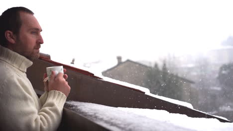 man drinking tea and playing with snowflakes on a balcony while is snowing