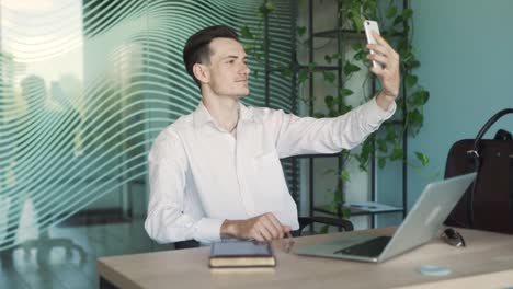 portrait of a handsome, young, stylish man in business attire, sitting at a desk in a modern office, taking a selfie with his smartphone