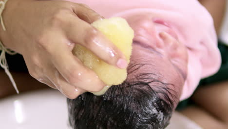 acunando la cabeza de un bebé con una mano mientras se enjuaga el cabello usando una esponja suave empapada en agua para lavar los residuos de jabón de la cabeza del bebé