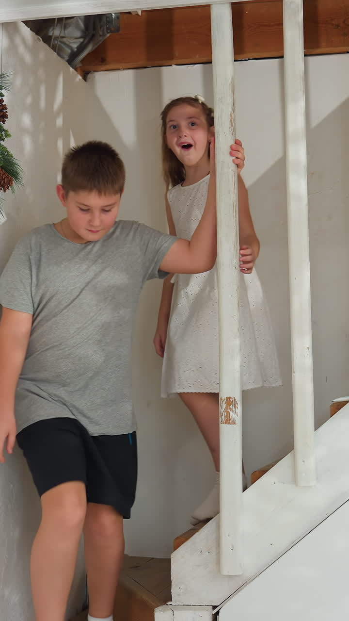white kids descending decorated staircase, brother leads with careful steps while sister beams under wreath shadow, candid family moment captures joyful energy and movement, cozy interior light